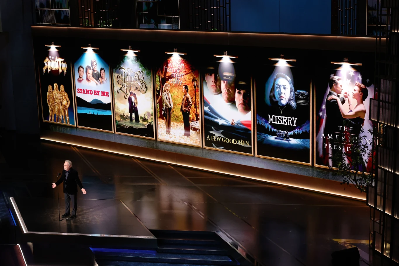 Duża scena z prezentacją oświetlonych, dużych plakatów kultowych filmów, takich jak 'Stand by Me', 'The Shawshank Redemption', 'The Green Mile', 'A Few Good Men', 'Misery' oraz 'The American President', z pojedynczą osobą stojącą przed mikrofonem z lew...
