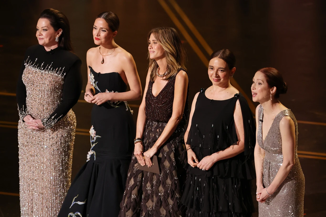 Pięć elegancko ubranych kobiet stoi w szeregu na scenie, każda w wieczorowej sukni, zwracając uwagę błyszczącymi detalami kreacji i skupionymi wyrazami twarzy, sugerując udział w uroczystym wydarzeniu.
