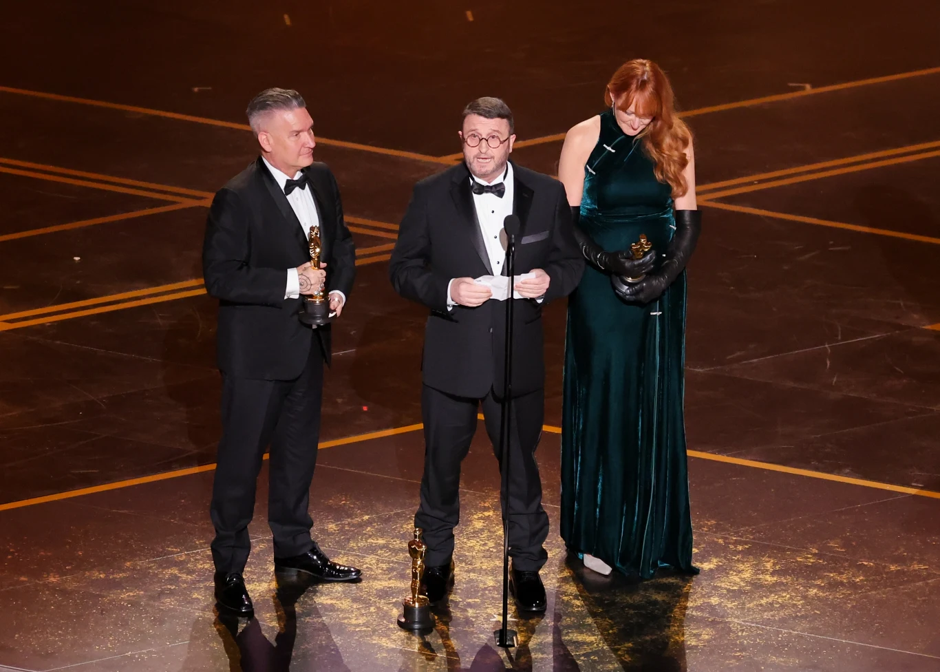 Trójka elegancko ubranych osób stoi na scenie z Oscarami w rękach, na środku znajduje się mężczyzna w smokingu mówiący do mikrofonu, po bokach towarzyszy mu inny mężczyzna oraz kobieta w zielonej, wieczorowej sukni.
