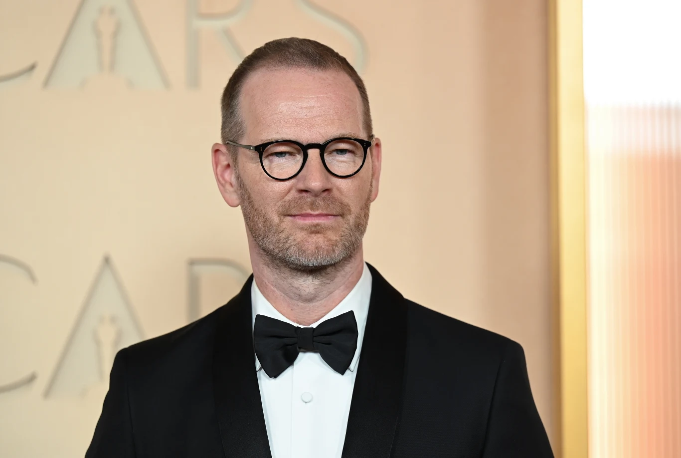 Mężczyzna w eleganckim czarnym smokingu z czarną muchą i białą koszulą, ma krótkie włosy, okulary o ciemnych oprawkach oraz lekki zarost, stoi na tle jasnej ścianki z widocznymi literami.