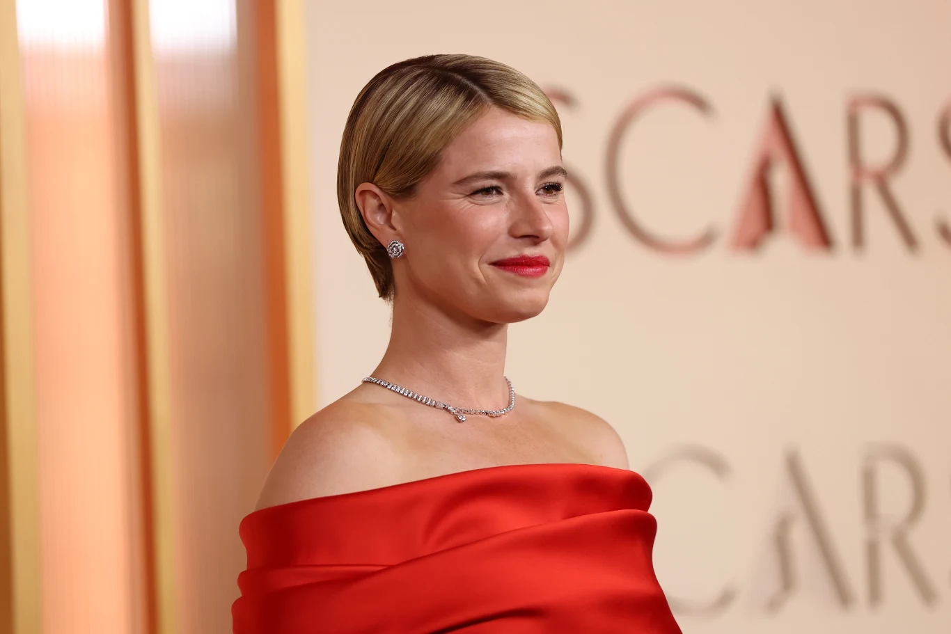 Jessie Buckley Jessie Buckley w czerwonej sukni, z krótkimi blond włosami, w eleganckiej biżuterii.