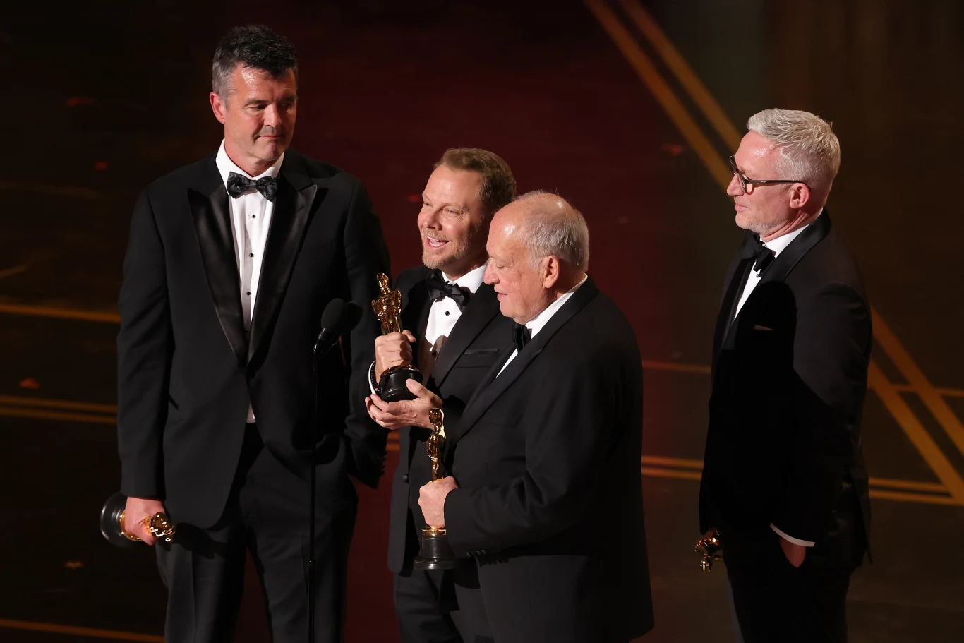 Czterech elegancko ubranych mężczyzn w smokingu stoi razem na scenie, dwóch z nich trzyma w dłoniach statuetki Oscara, wszyscy są uśmiechnięci i zwróceni w stronę mikrofonu.