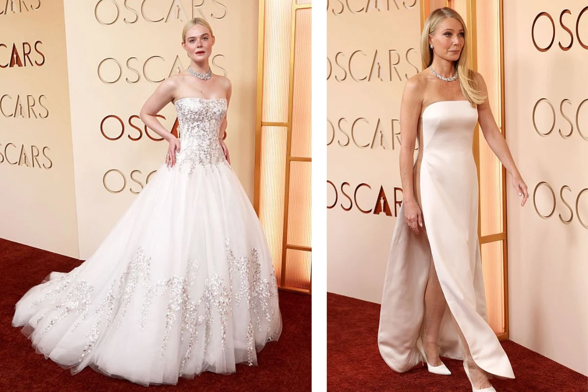 Dwie kobiety w eleganckich, jasnych sukniach stoją na czerwonym dywanie przed ścianą z napisem 'OSCARS'; pierwsza ma na sobie rozkloszowaną suknię z haftami, druga długą, dopasowaną kreację bez ramiączek.