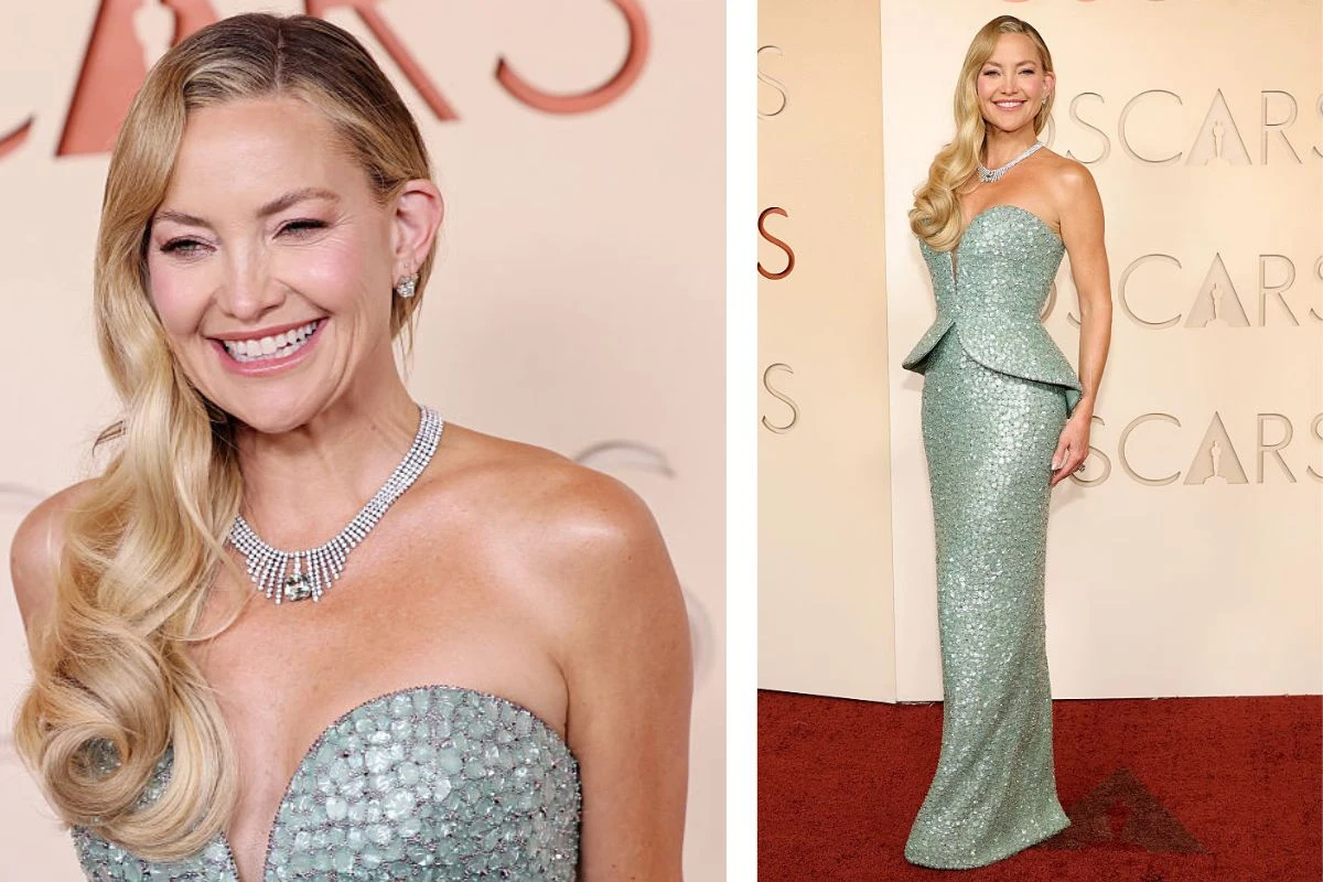 Kobieta w długiej, błyszczącej, jasnozielonej sukni z gorsetem ozdobionym cekinami, z długimi, falowanymi blond włosami opadającymi na jedno ramię, uśmiechnięta, z elegancką biżuterią na szyi, pozuje na czerwonym dywanie na tle ściany z napisem OSCARS.