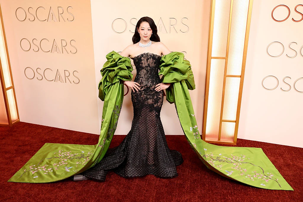 Kobieta w efektownej, czarnej sukni z dużymi, zielonymi, ozdobnymi rękawami, ozdobiona biżuterią, pozuje na czerwonym dywanie przed ścianą z napisem „OSCARS”.