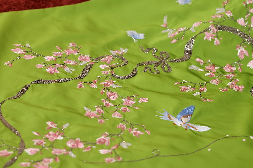 Jasnozielony materiał ozdobiony haftem przedstawiającym gałęzie drzewa z różowymi kwiatami oraz niebieskie ptaki w locie.