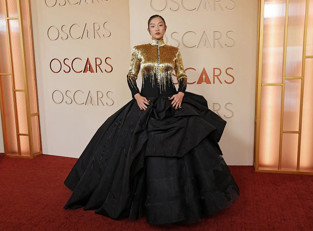 Kobieta w efektownej, złoto-czarnej sukni z rozłożystym dołem pozuje na czerwonym dywanie podczas gali Oscarów przed ścianką z logotypem imprezy.