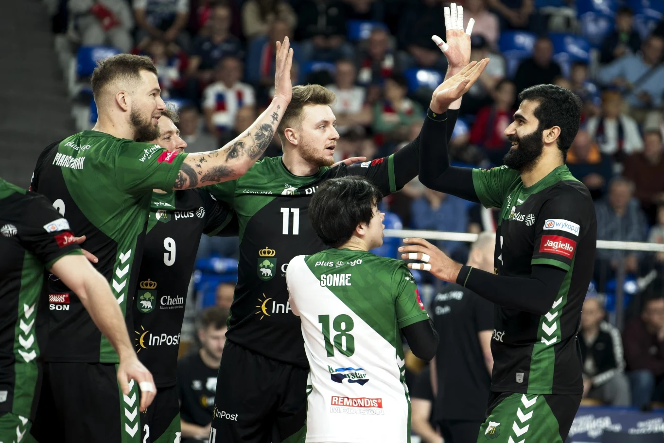 Grupa siatkarzy w zielono-czarnych strojach świętuje udany punkt, wymieniając piątki i okazując radość na boisku podczas meczu, na trybunach widoczni kibice.