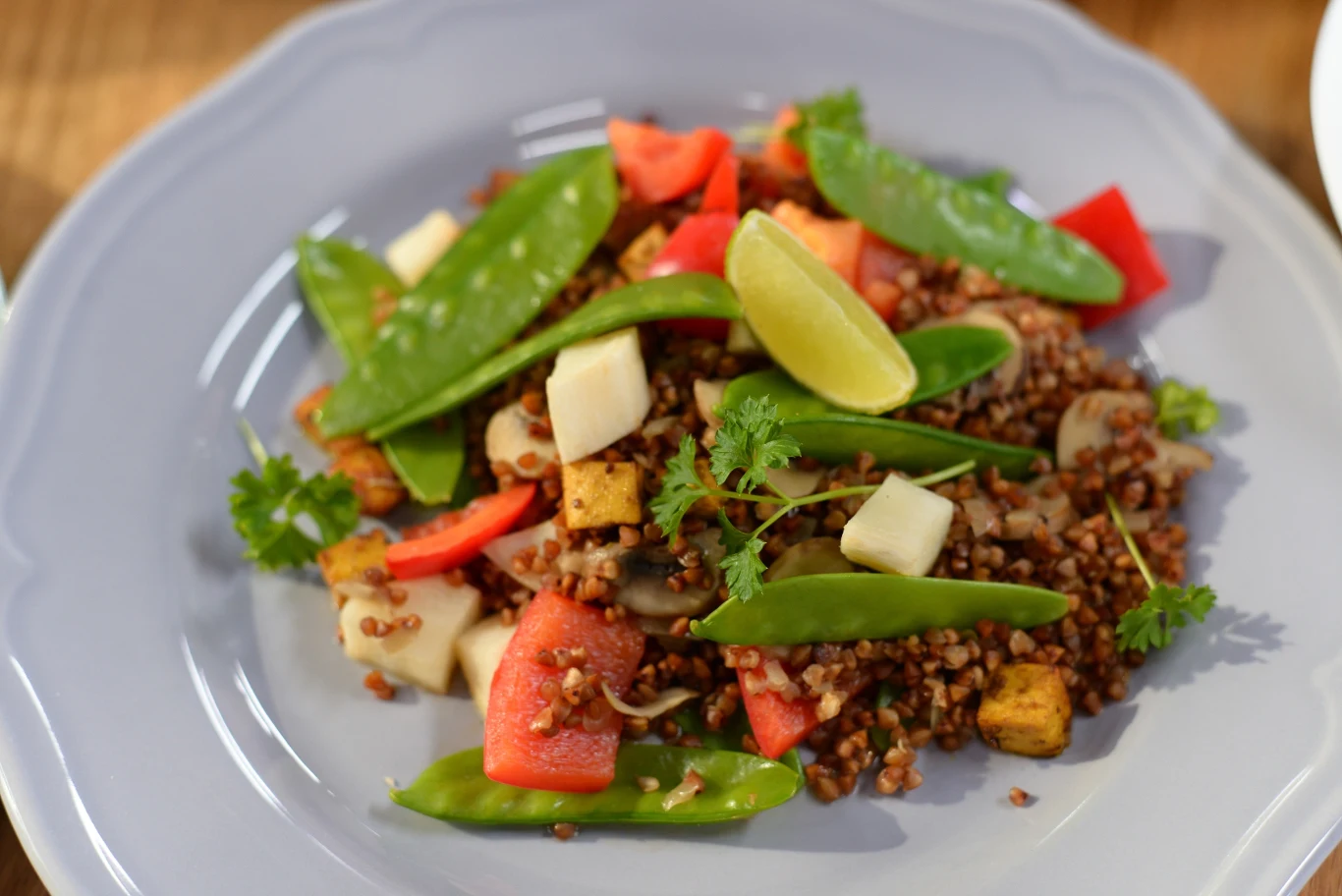 Kompozycja kaszy gryczanej z kawałkami tofu, groszkiem cukrowym, papryką czerwoną i plasterkami białego sera, udekorowana świeżą kolendrą oraz ćwiartką limonki.