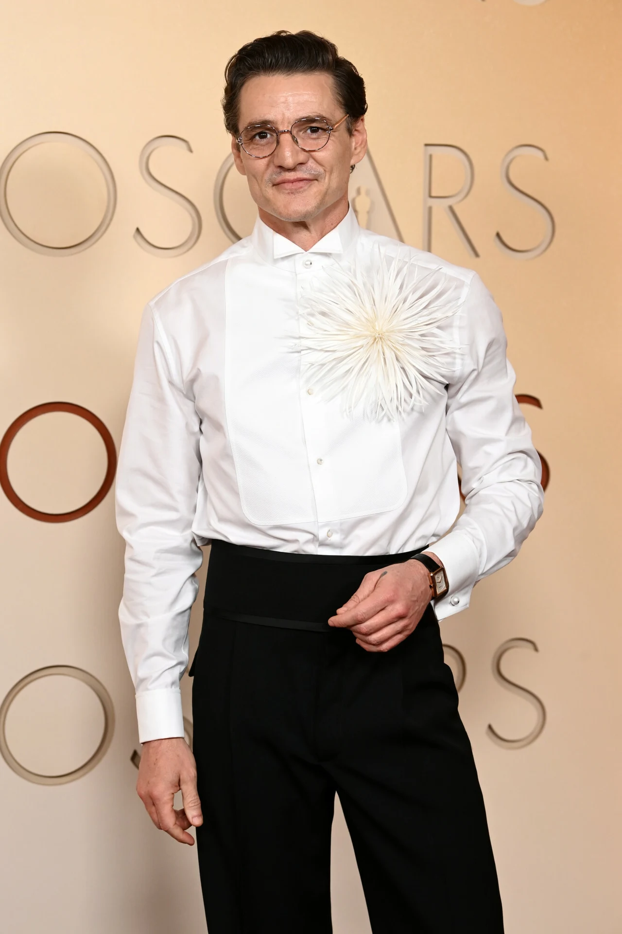 Pedro Pascal Pedro Pascal w eleganckim stroju, ubrany w białą koszulę z ozdobną aplikacją w postaci dużego, białego kwiatu na piersi oraz czarne, wysokie spodnie na galo Oscarów 2026.