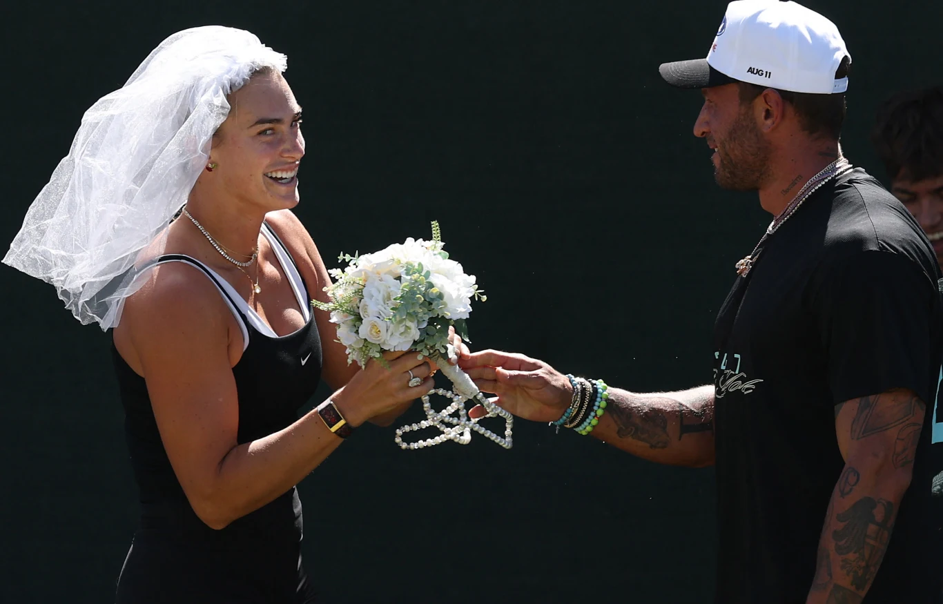 Uśmiechnięta kobieta w welonie trzyma bukiet kwiatów i symboliczną tiarę, odbierając je od mężczyzny w sportowej czapce na tle ciemnego tła.