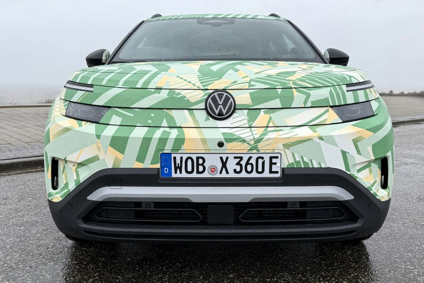 Nowoczesny samochód elektryczny marki Volkswagen z jasnym, zielono-żółtym wzorem maskującym na karoserii, widoczny z przodu na tle pochmurnego nieba; centralnie umieszczony emblemat VW oraz niemieckie tablice rejestracyjne.