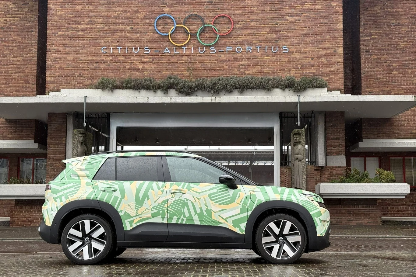 Nowoczesny samochód terenowy w zielono-żółtym kamuflażu zaparkowany przed budynkiem z cegły, na którym widnieją olimpijskie koła oraz motto 'Citius-Altius-Fortius'.