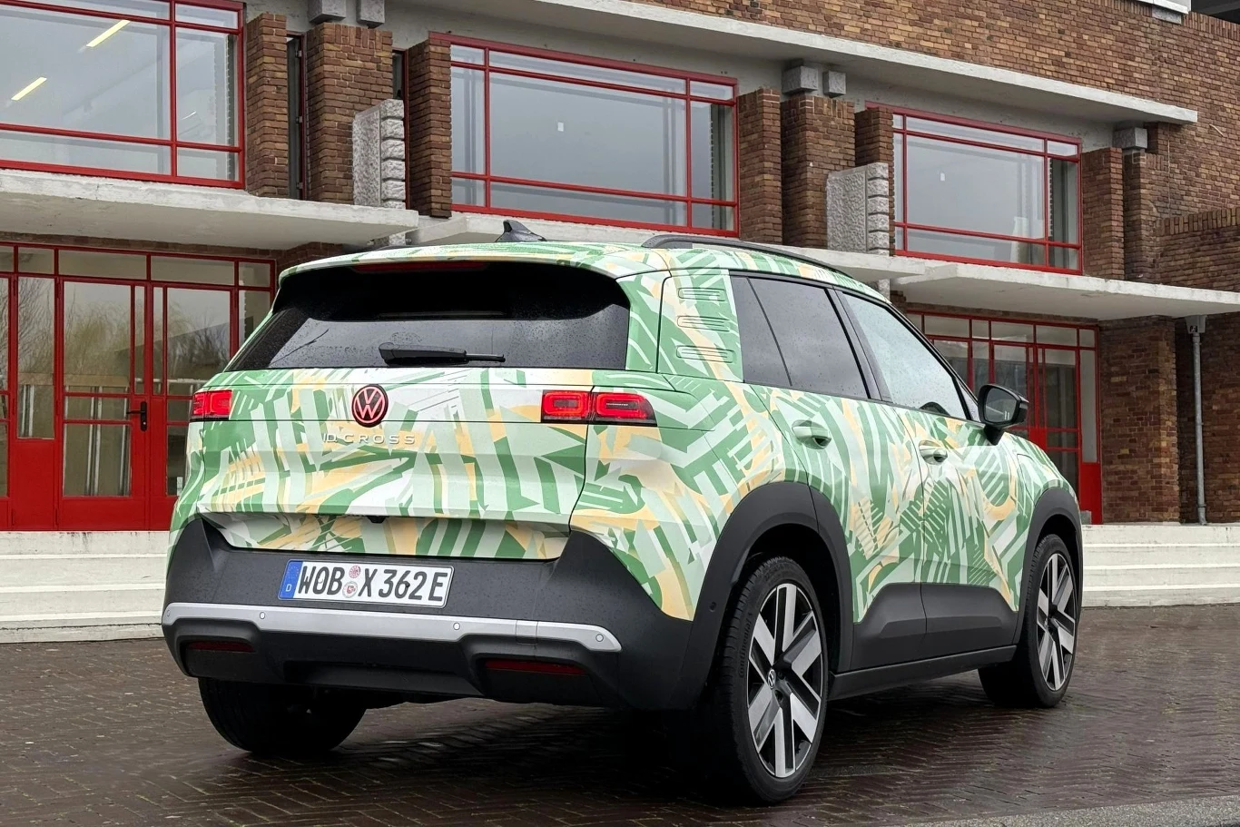 Samochód marki Volkswagen w zielonym kamuflażu z roślinnym wzorem, zaparkowany na brukowanej powierzchni przed nowoczesnym budynkiem z czerwonymi oknami i drzwiami.