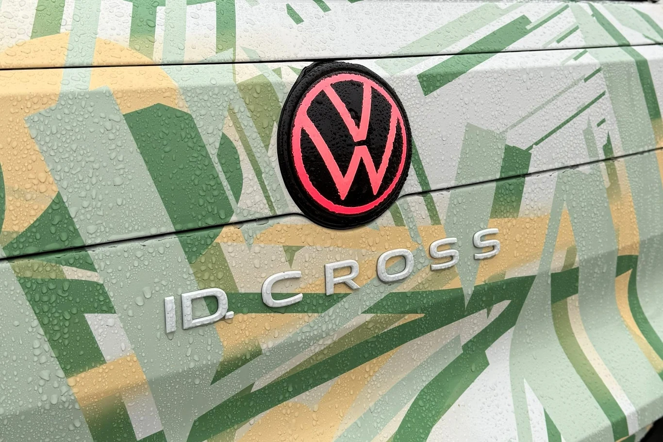 Logo Volkswagena w neonowym różowym kolorze oraz napis 'ID. CROSS' na tle karoserii pokrytej zielono-żółtym, geometrycznym wzorem z kroplami wody.