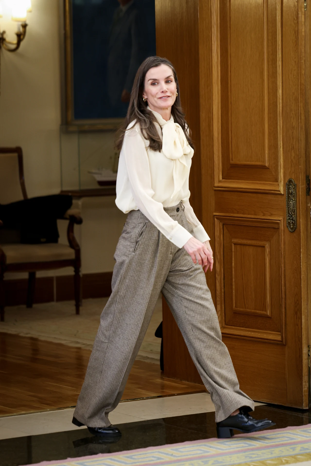 Kobieta z długimi, ciemnymi włosami ubrana w elegancką, kremową bluzkę z kokardą przy szyi oraz szerokie, szare spodnie w kant, idzie przez jasny, przestronny pokój z drewnianymi drzwiami w tle, na nogach ma czarne buty na płaskiej podeszwie.