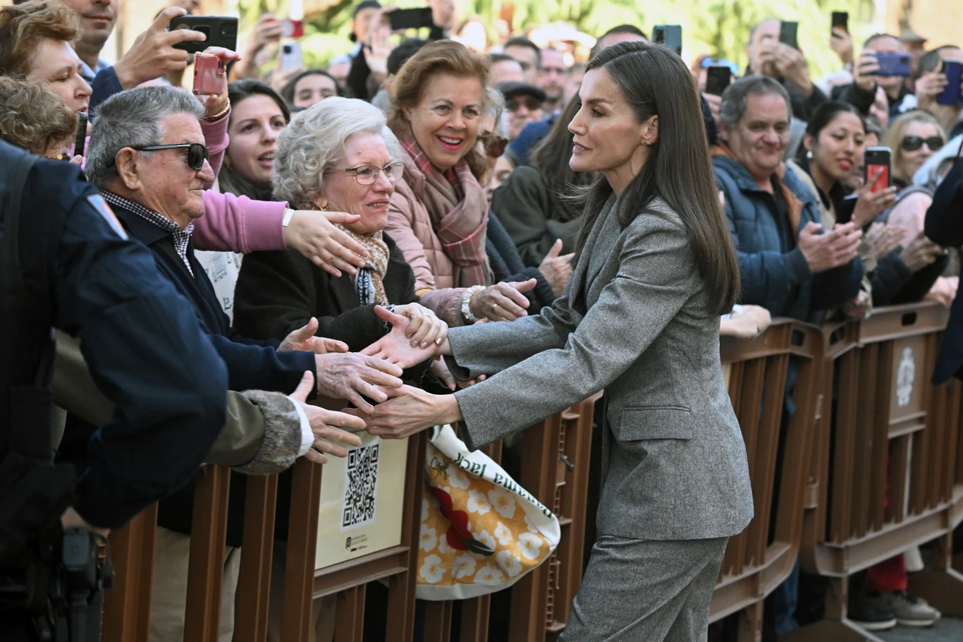 Elegancko ubrana kobieta w szarym garniturze nawiązuje kontakt z tłumem, ściskając dłonie i uśmiechając się do ludzi stojących za barierkami; wielu z nich wyciąga ręce, robi zdjęcia i okazuje entuzjazm.