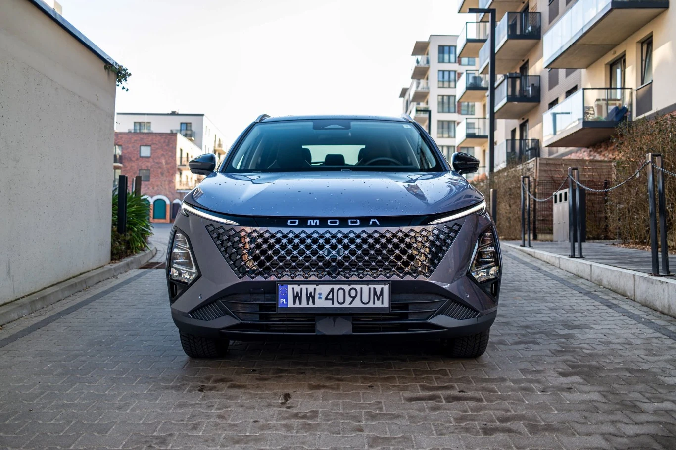 Nowoczesny SUV Omoda z charakterystycznym dużym grillem i futurystycznymi reflektorami, zaparkowany na brukowanej alejce w otoczeniu nowoczesnych budynków mieszkalnych.