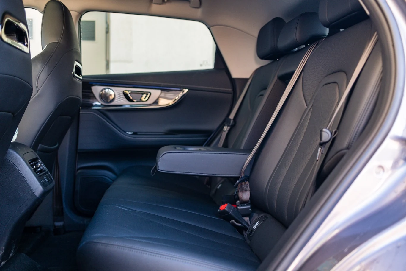 Eleganckie, czarne skórzane siedzenia tylnego rzędu samochodu z opuszczonym podłokietnikiem i zapiętymi pasami bezpieczeństwa, dobrze widoczne detale wykończenia drzwi oraz otwarte okno po prawej stronie.