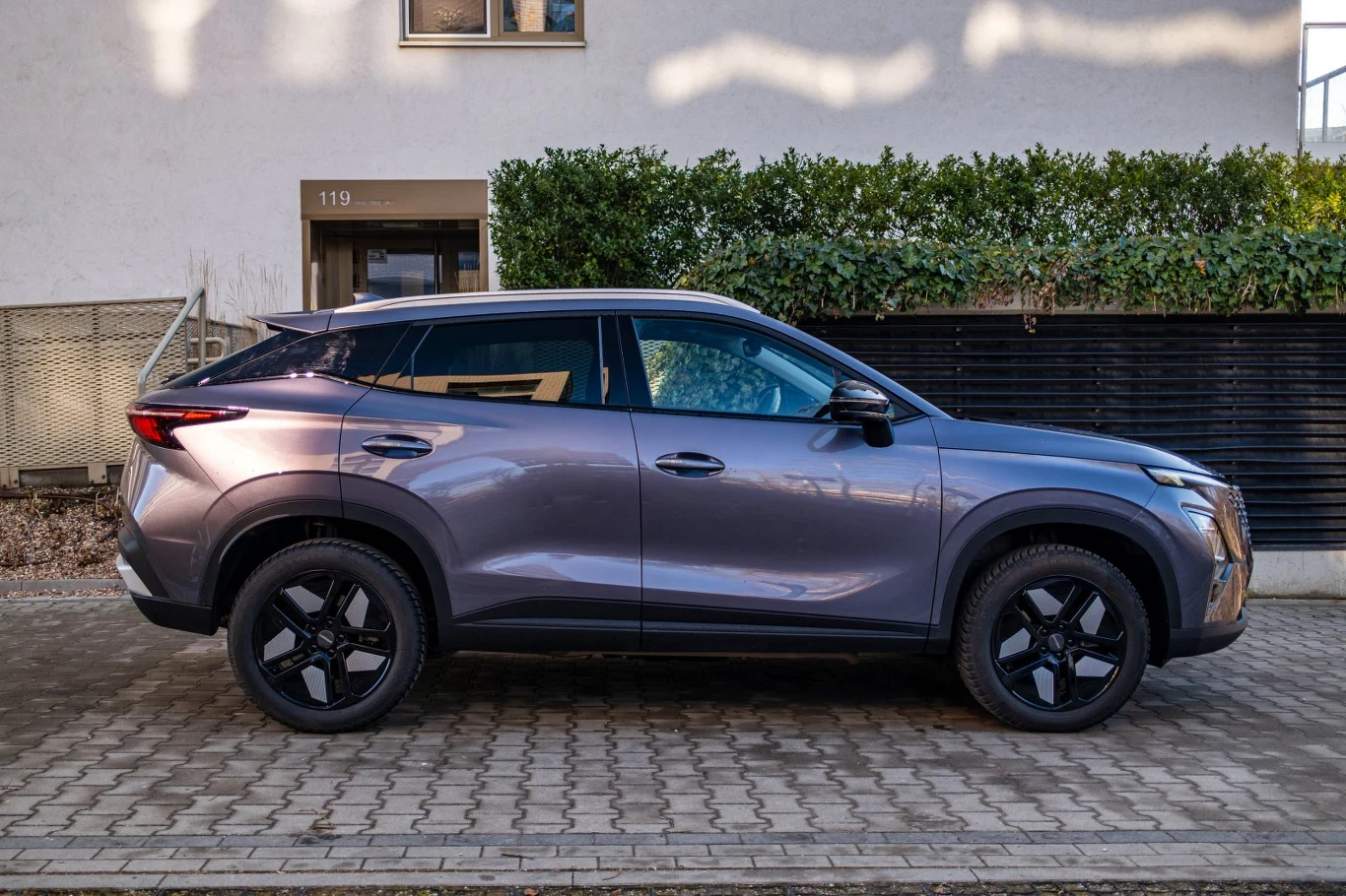 fioletowy SUV o dynamicznej sylwetce i czarnych felgach zaparkowany przy krawężniku, w tle zielony żywopłot i jasny budynek z oknem oraz numerem 119 nad drzwiami