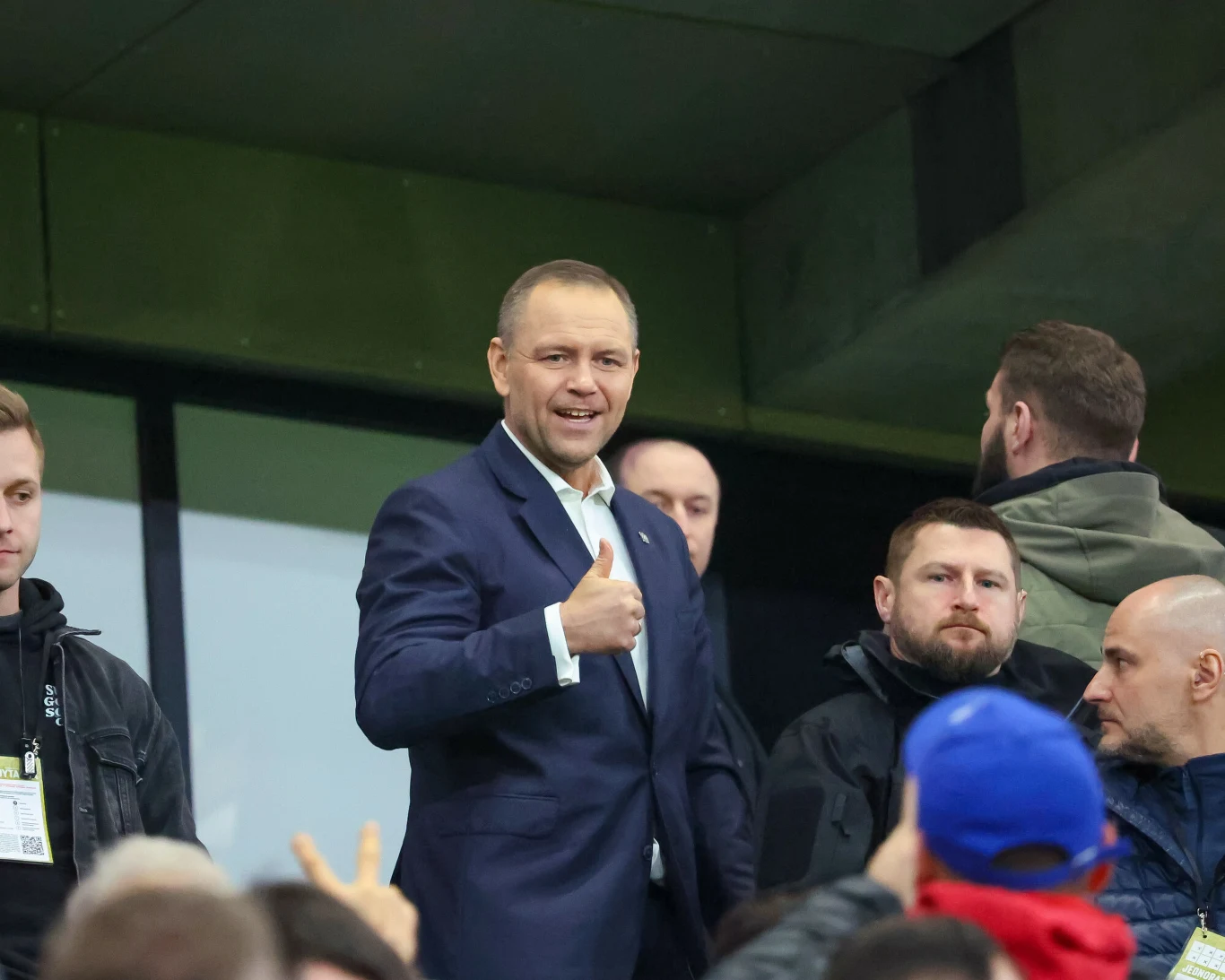 Mężczyzna w garniturze uśmiecha się i pokazuje gest kciuka w górę, otoczony przez kilku innych ludzi na trybunach stadionu.