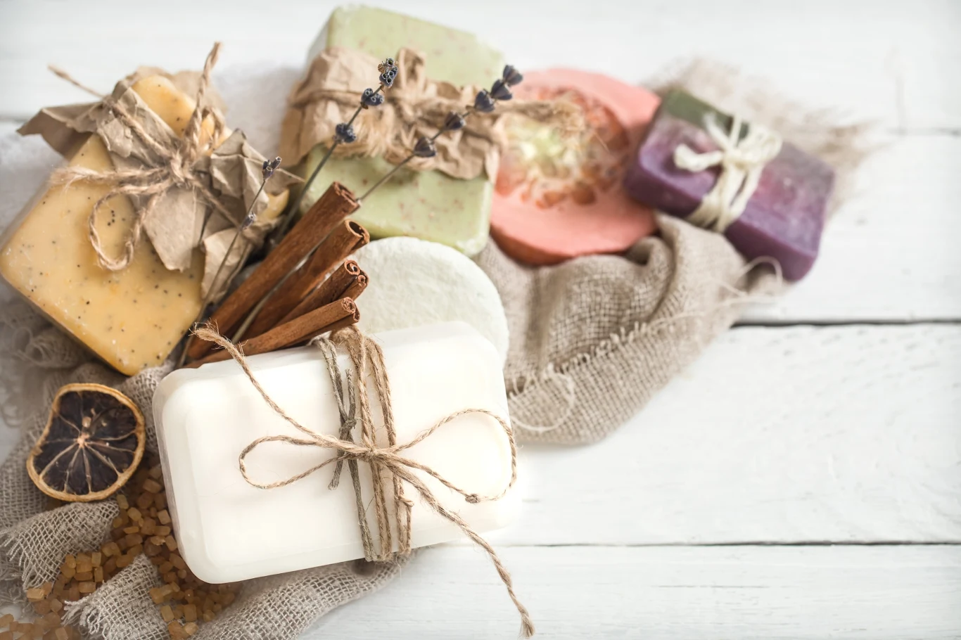 kolorowe, ręcznie robione mydła przewiązane sznurkiem jutowym, położone na lnianej tkaninie obok suszonego plastra cytryny, lasek cynamonu i suszonego kwiatu lawendy