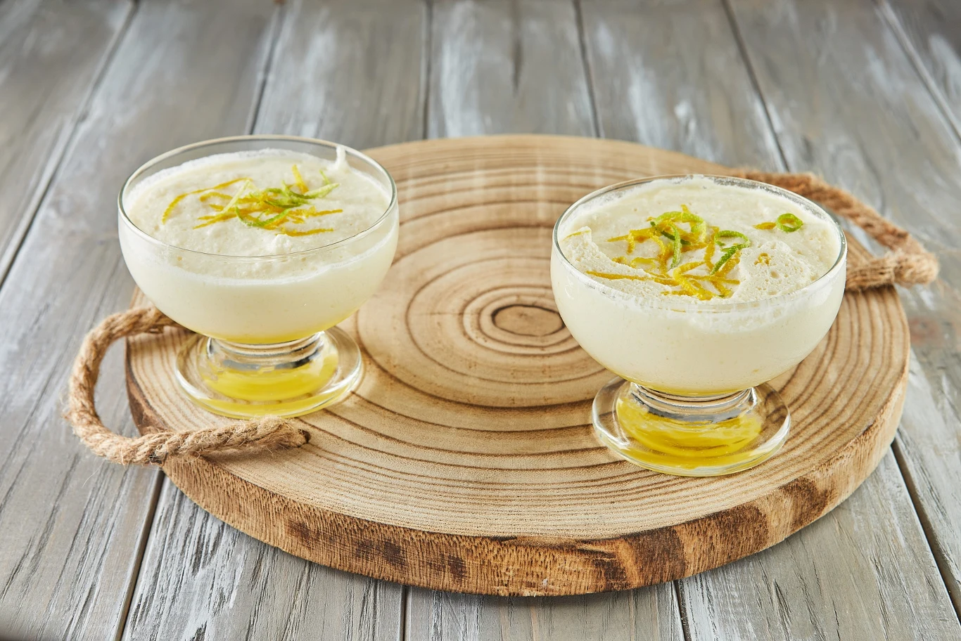 Dwie szklane pucharki wypełnione kremowym deserem udekorowanym startą skórką cytrynową, ustawione na okrągłej drewnianej desce z uchwytami ze sznura, całość na tle jasnego, drewnianego stołu.
