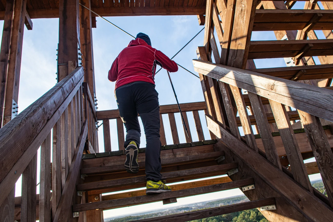 osoba w sportowym ubraniu wspinająca się po drewnianych schodach wieży widokowej, energiczny ruch, otoczenie wykonane z drewna, elementy zabezpieczające w postaci lin oraz widok na krajobraz za barierkami