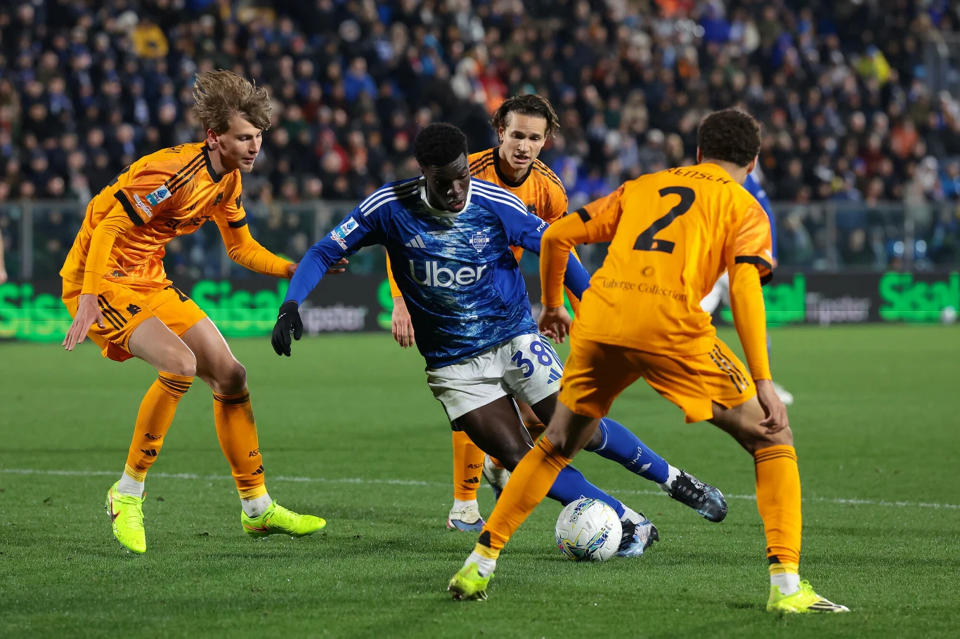 Jan Ziółkowski w niebieskiej koszulce walczy o piłkę z trzema rywalami w pomarańczowych strojach na stadionie.