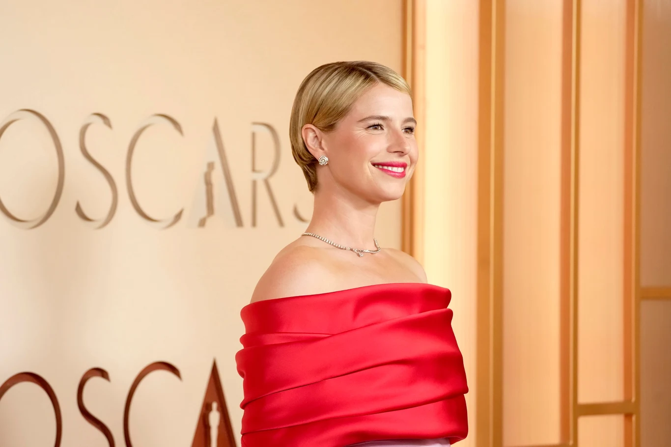 elegancko ubrana kobieta z krótkimi blond włosami pozuje na tle napisu 'OSCARS', ubrana w efektowną czerwoną suknię odsłaniającą ramiona, z delikatną biżuterią i wyrazistym uśmiechem