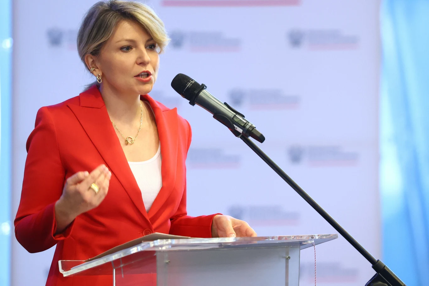 Kobieta w czerwonym żakiecie przemawia przy mównicy, gestykulując ręką i patrząc przed siebie, a przy jej twarzy znajduje się mikrofon na statywie.