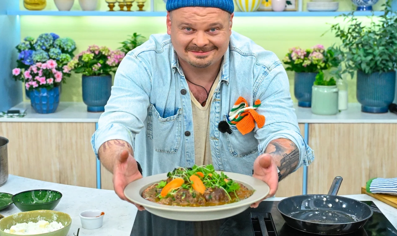 mężczyzna w niebieskiej czapce i dżinsowej kurtce trzyma talerz z daniem udekorowanym świeżymi ziołami i warzywami, stojąc w nowoczesnej kuchni z ozdobnymi roślinami doniczkowymi w tle