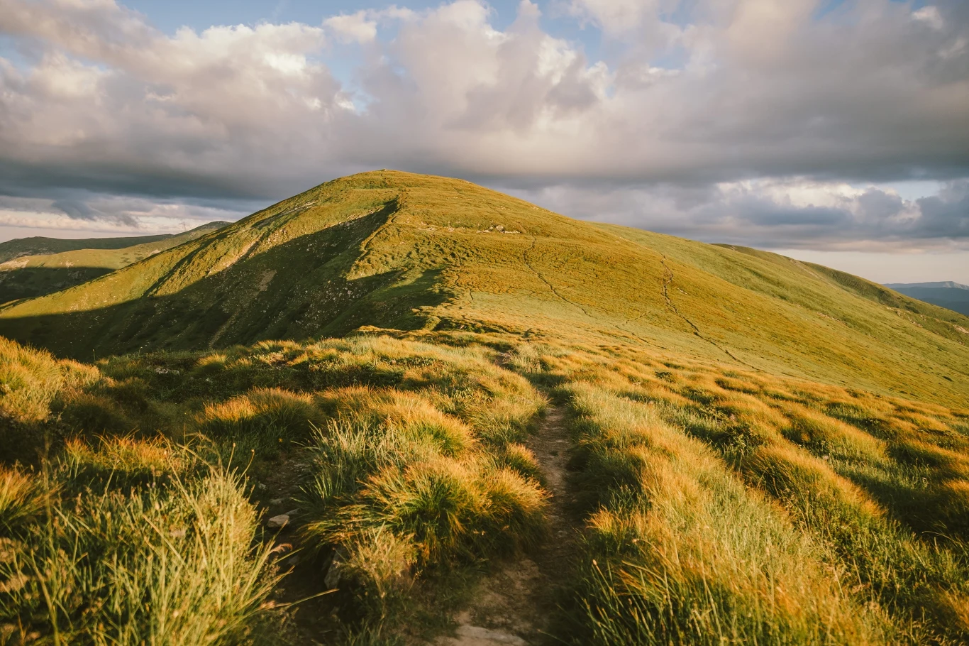 Połoniny bieszczadzkie zielone wzgórze pokryte trawą, oświetlone promieniami zachodzącego słońca, z wyraźnie widoczną ścieżką prowadzącą na szczyt pod pochmurnym niebem