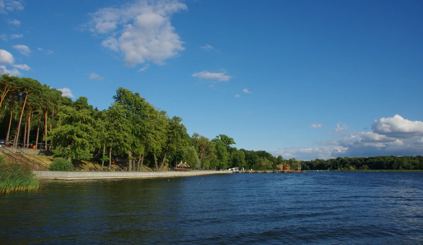 spokojna tafla jeziora otoczona zielonym lasem, po lewej stronie wąska promenada biegnąca wzdłuż brzegu, na horyzoncie błękitne niebo z kilkoma białymi chmurami