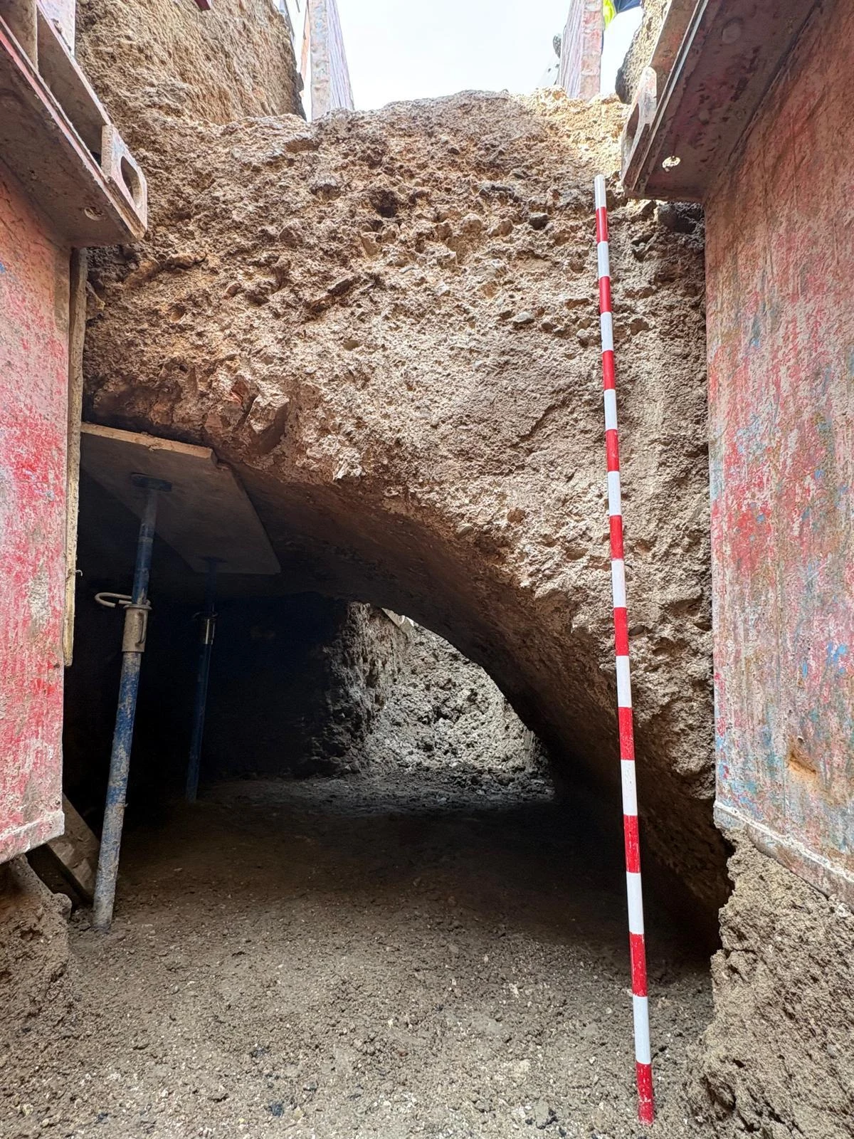 Fragment odkrytego mostu łukowata struktura ziemna w wykopie podparta metalowymi podporami, obok której ustawiona jest czerwonobiała miarka geodezyjna; widoczna surowa powierzchnia ziemi bez wyraźnych śladów obecności innych elementów