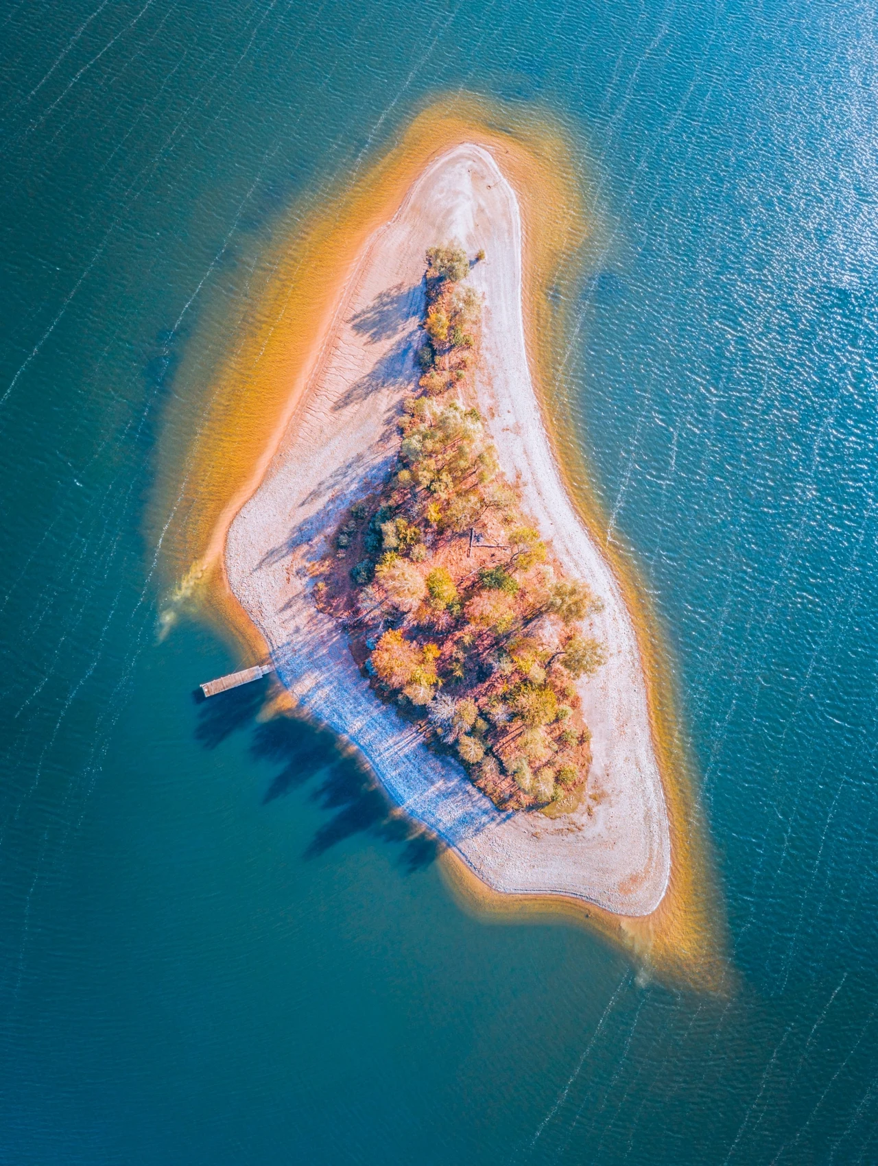 Mała wyspa na jeziorze Solińskim, Polańczyk Mała, piaszczysta wyspa otoczona błękitną wodą, porośnięta kolorowymi drzewami w jesiennych barwach, z drewnianym pomostem sięgającym w stronę jeziora.
