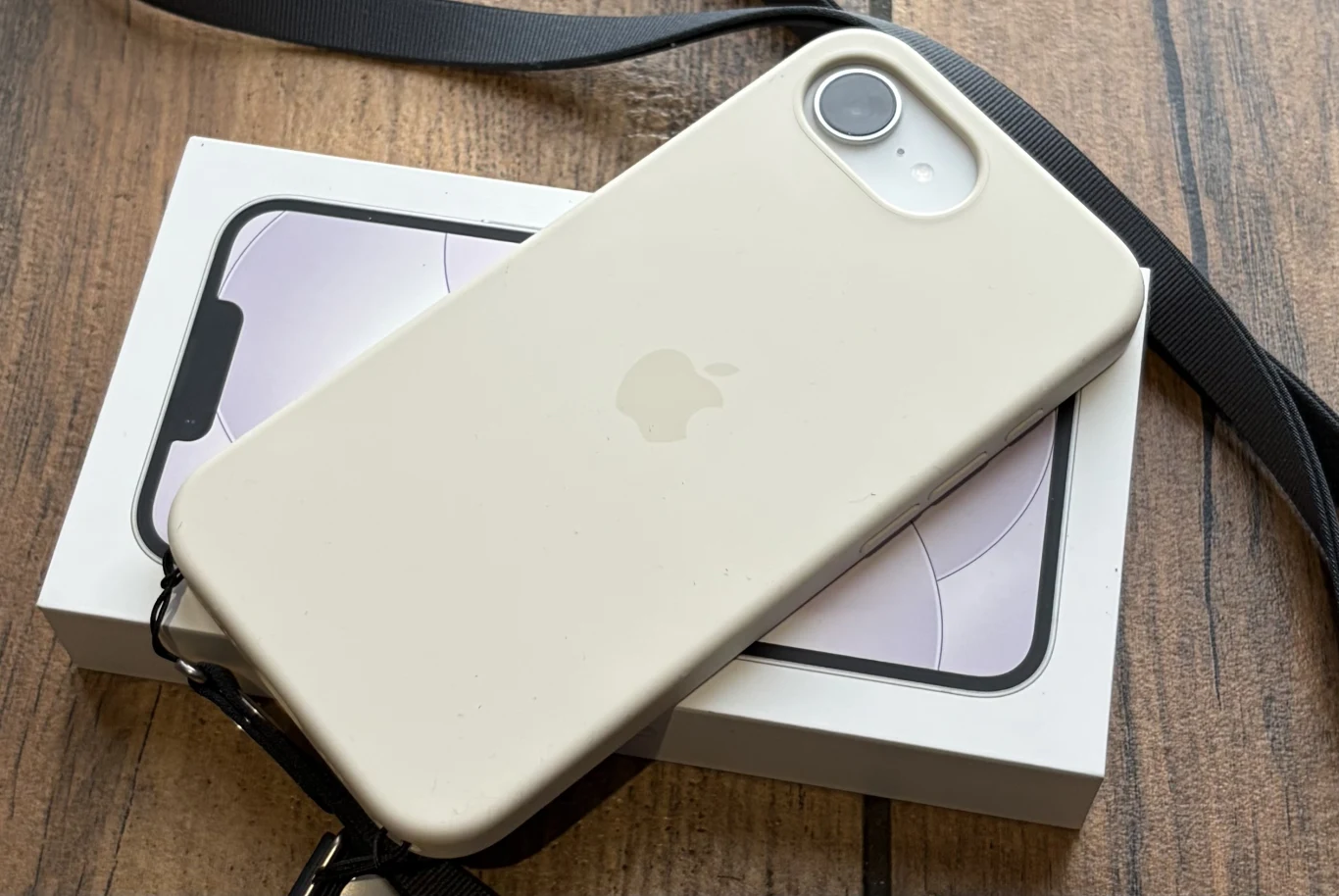 Smartfon iPhone 17e - pierwsze wrażenia. Czy warto go kupić? iPhone 17e w silikonowym etui ze smyczą leży na opakowaniu, całość na drewnianej powierzchni