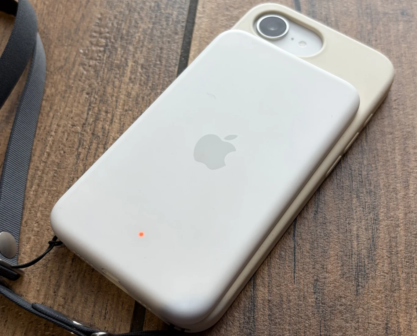 iPhone 17e działa w połączeniu z zewnętrzną baterią Apple dla iPhone'a Air. Biała przenośna ładowarka MagSafe przyczepiona do tyłu białego iPhone’a, leżących na drewnianej powierzchni; na ładowarce widoczne subtelne logo Apple i mała czerwona dioda sygnalizująca pracę urządzenia.