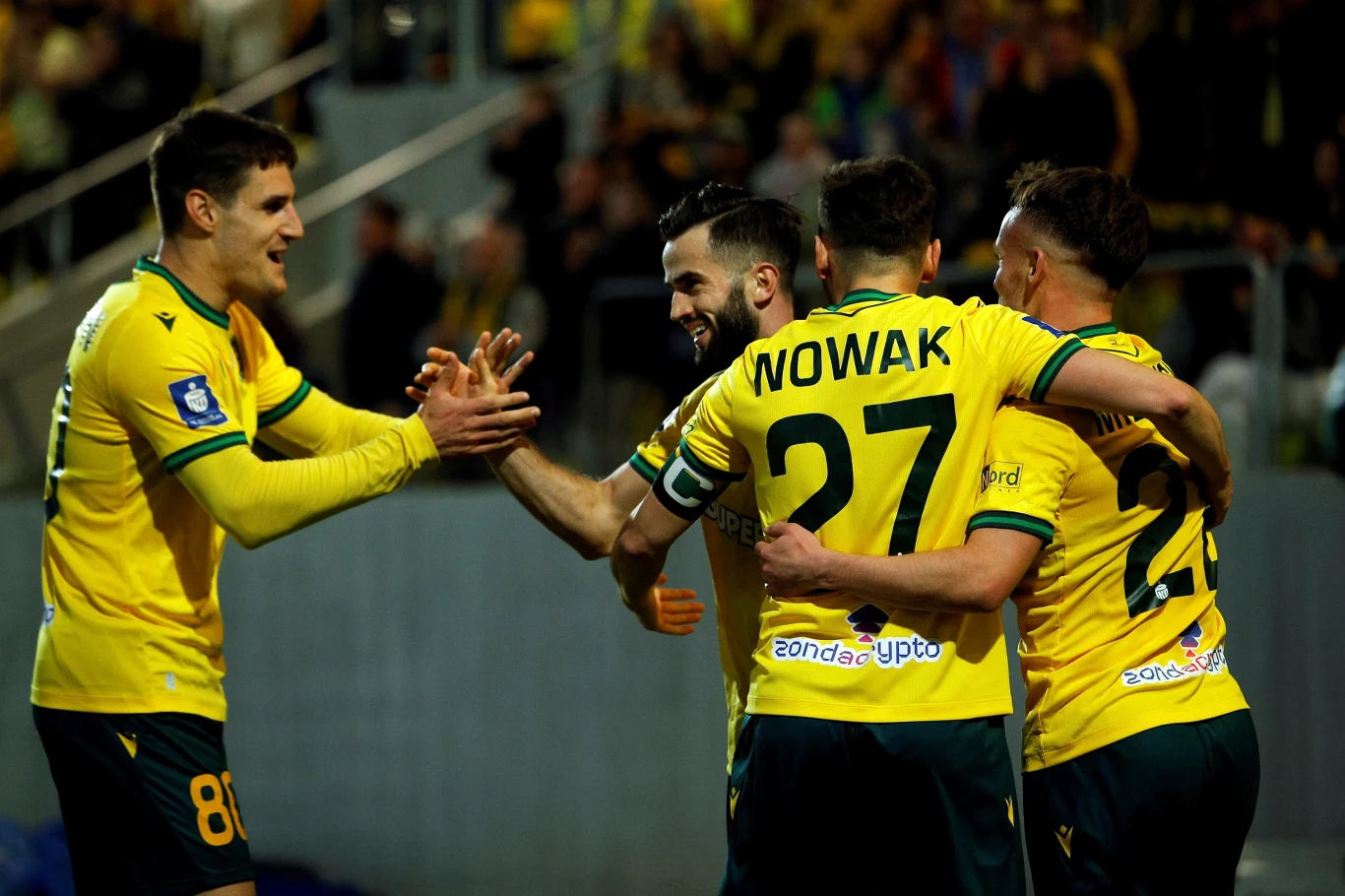 Czterech piłkarzy w żółto-zielonych strojach świętuje zdobycie bramki, przybijając sobie piątki i obejmując się na tle stadionu wypełnionego kibicami.