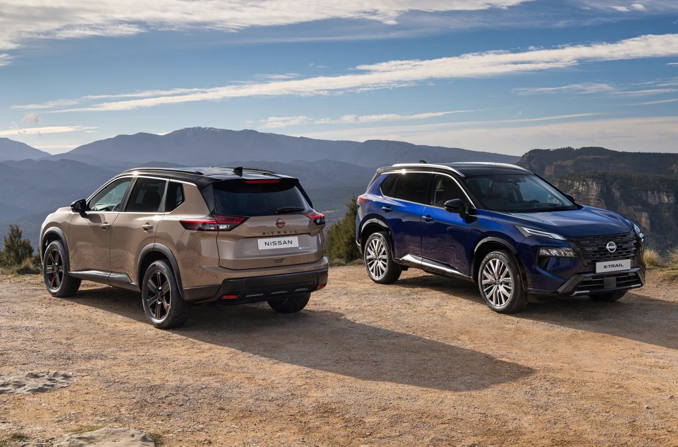 Dwa nowoczesne SUV-y Nissan X-Trail zaparkowane na otwartym terenie z widokiem na góry, jeden w kolorze beżowym od tyłu, drugi w niebieskim od przodu, tworzą kontrastową kompozycję na tle naturalnego krajobrazu.