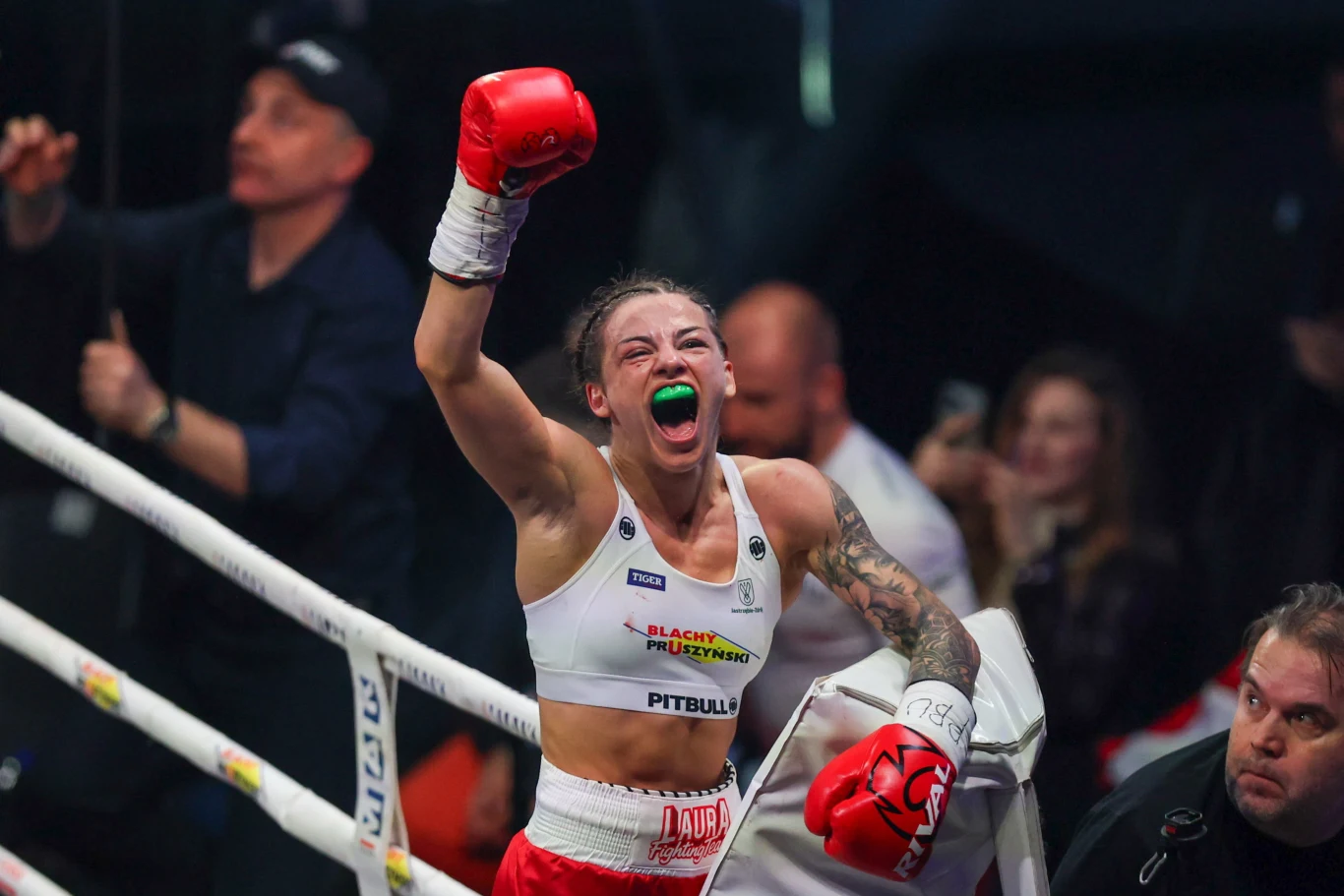 Bokserka w ringu z uniesioną w geście triumfu ręką, z wyrazem radości na twarzy oraz ochraniaczem na zęby, otoczona kibicami i członkami zespołu.