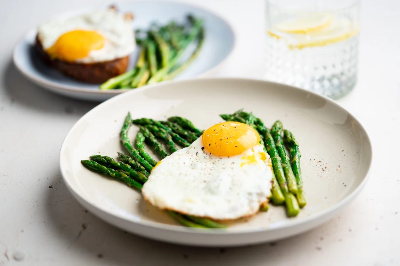 Porcja zielonych szparagów ułożona na białym talerzu, na nich smażone jajko sadzone z płynnym żółtkiem, w tle drugi talerz z podobnym daniem oraz szklanka wody z plasterkiem cytryny.