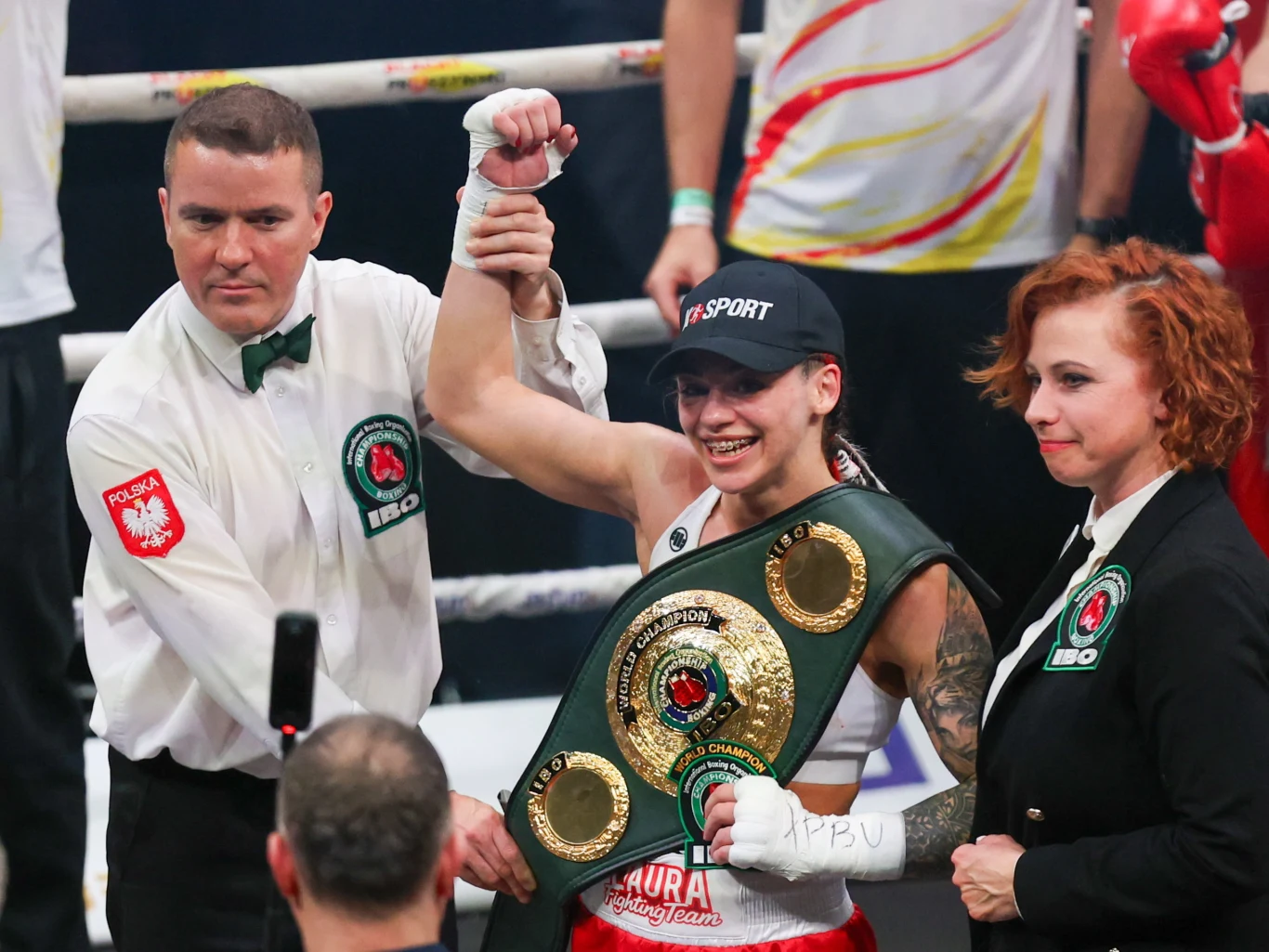 Zwyciężczyni walki bokserskiej trzyma podniesioną przez sędziego rękę, szeroko się uśmiecha i ma na sobie czapkę oraz pas mistrzowski IBO na ramionach; obok stoi kobieta z identyfikatorem organizatora.