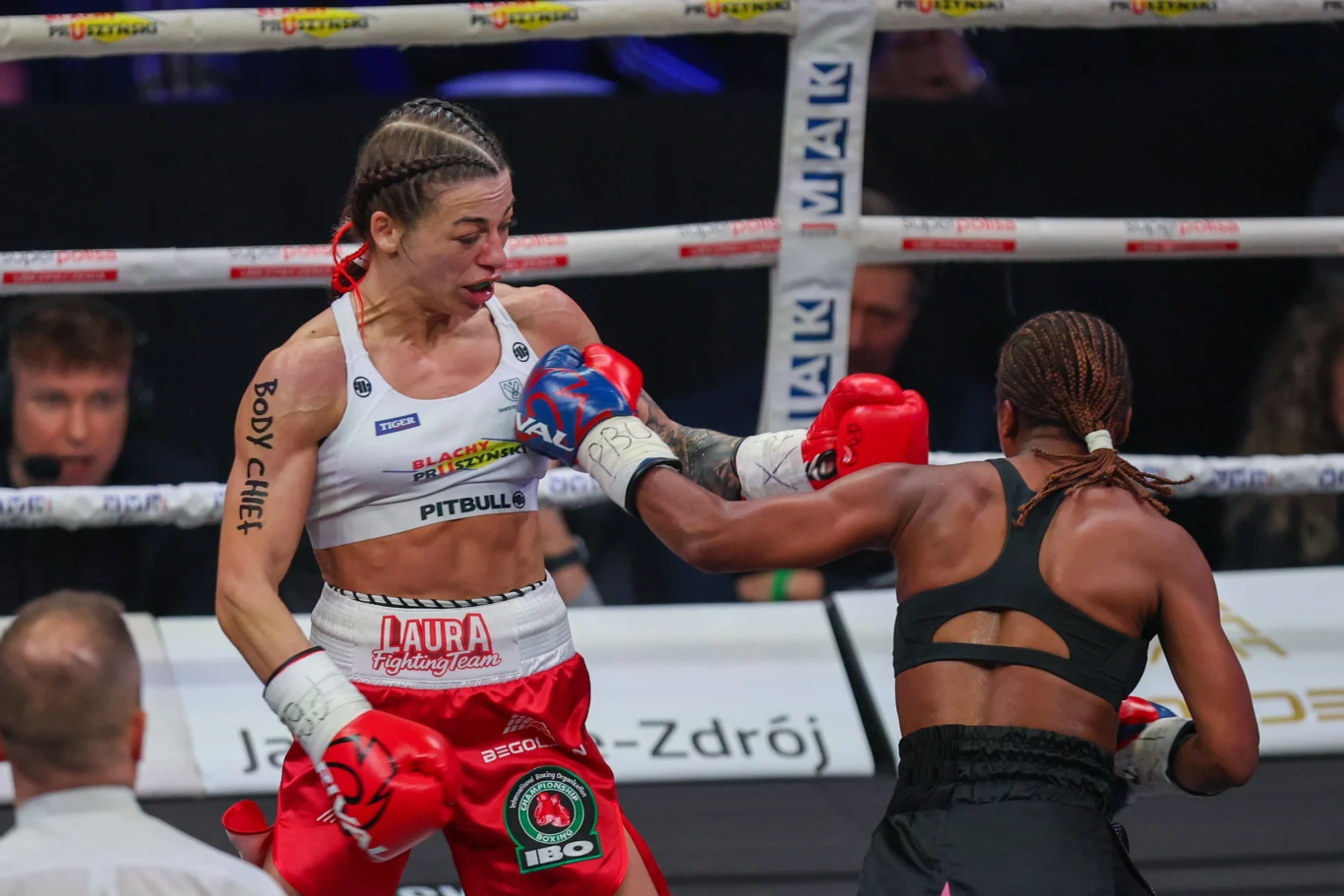 dwie kobiety walczące w ringu bokserskim, jedna z nich w czerwonych spodenkach wyprowadza cios prawą ręką w kierunku głowy przeciwniczki w czarnym stroju