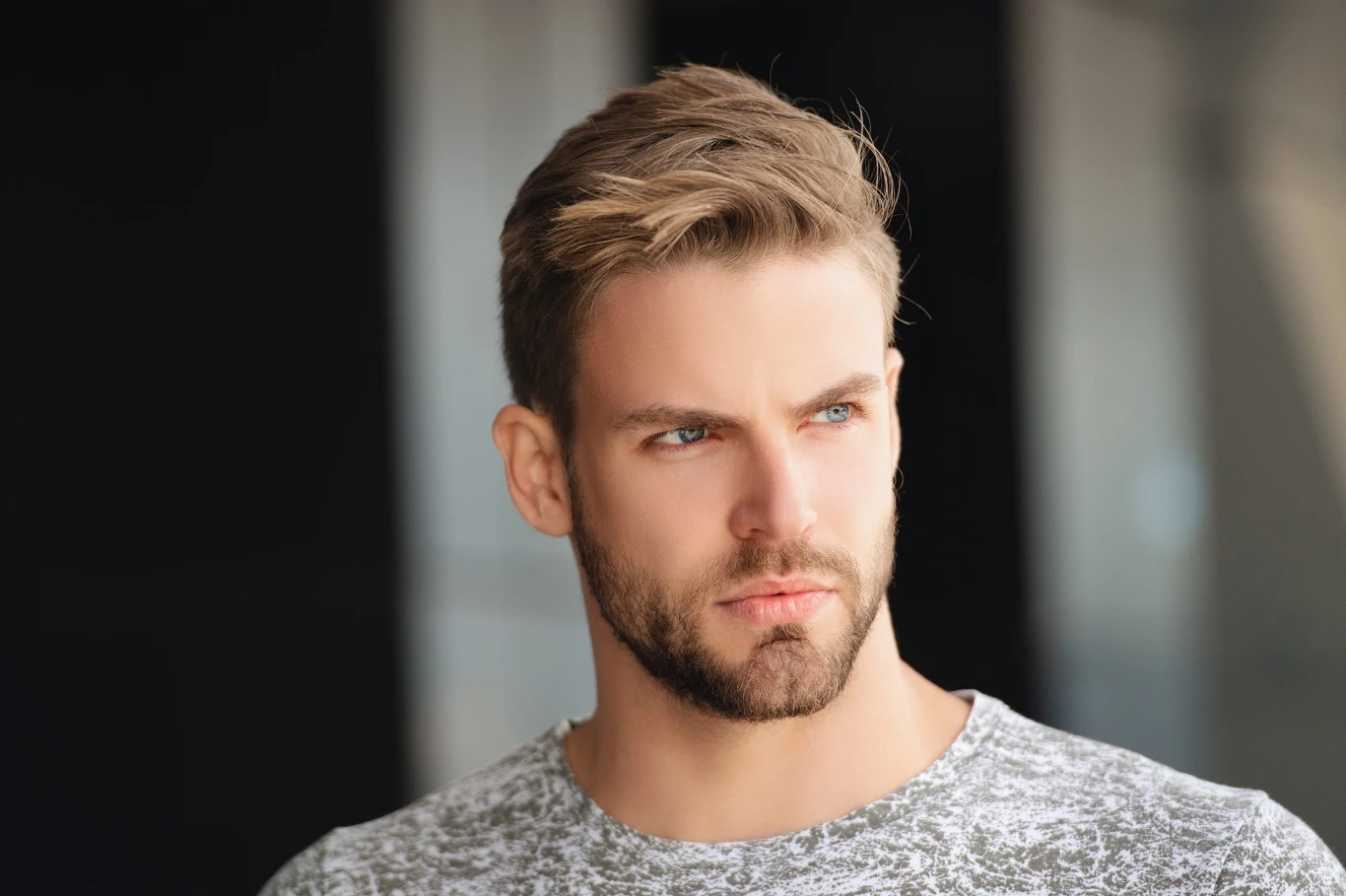 Młody mężczyzna z krótką blond fryzurą i brodą, ubrany w jasnoszarą koszulkę, spogląda poważnie w bok na neutralnym tle.