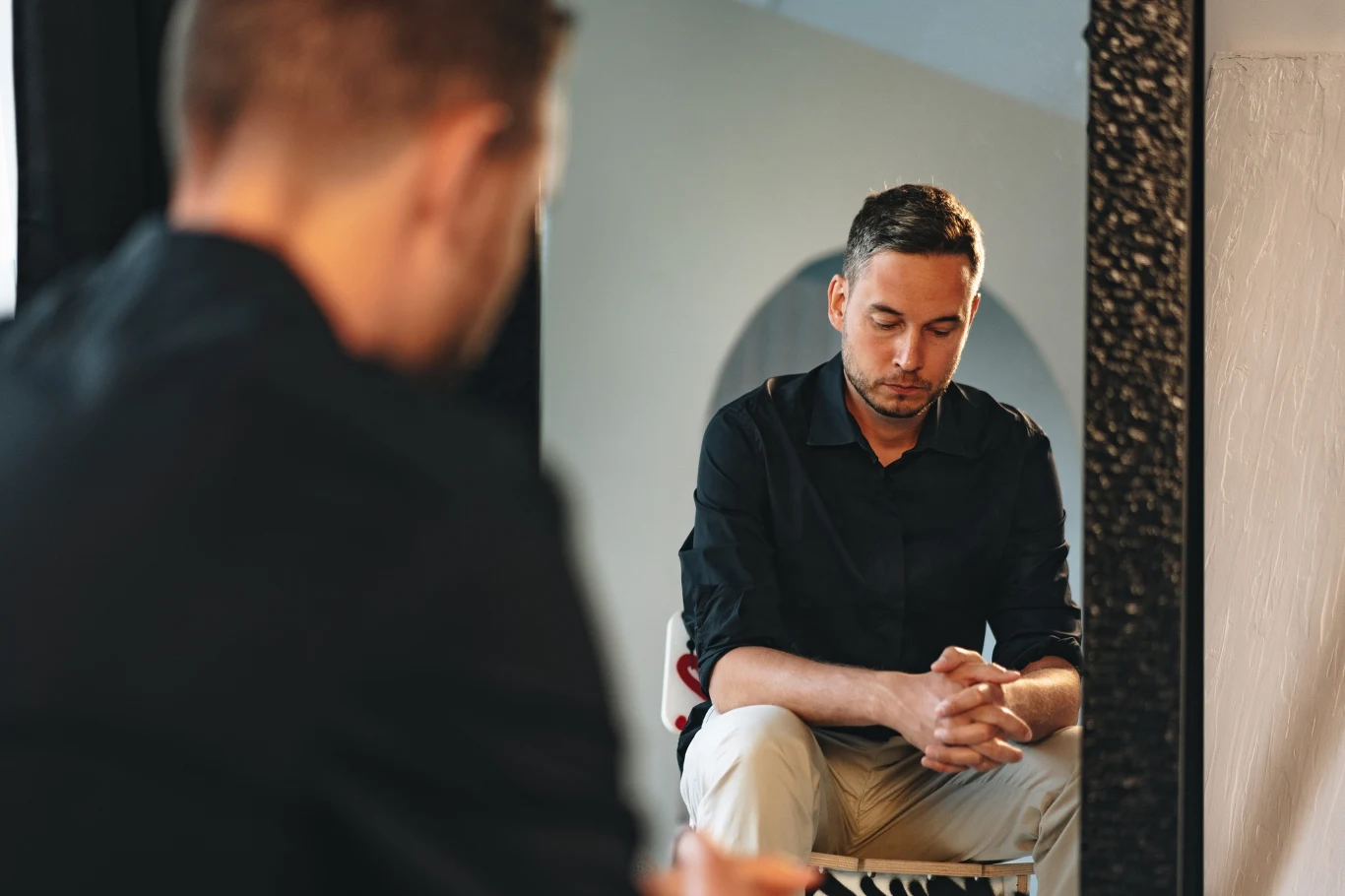 Mężczyzna w czarnej koszuli siedzi na krześle, patrząc w dół z poważnym wyrazem twarzy, jego dłonie są splecione, a w lustrze widoczne jest jego odbicie.