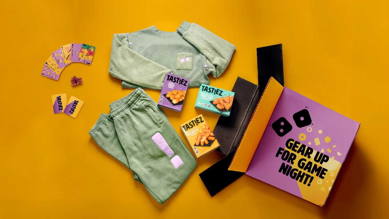 Zestaw zielonej bluzy i spodni dresowych z naszywkami, kolorowe przekąski Tastez, otwarte pudełko z napisem 'Gear up for game night!', kilka kart i kość do gry na intensywnie żółtym tle.