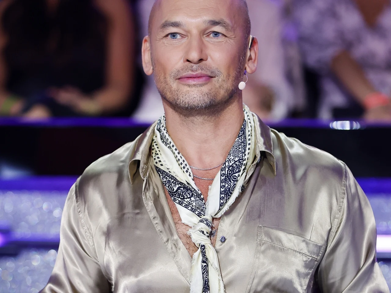 Mężczyzna w jedwabnej, błyszczącej koszuli i wzorzystej apaszce, z mikrofonem przy twarzy, siedzi w jasnym, oświetlonym studiu telewizyjnym
