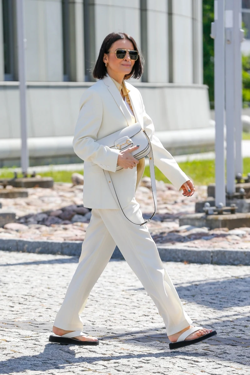 kobieta w eleganckim, jasnym garniturze i sandałach spaceruje chodnikiem, trzymając w ręku białą torebkę na pasku, na twarzy okulary przeciwsłoneczne, krótka ciemna fryzura