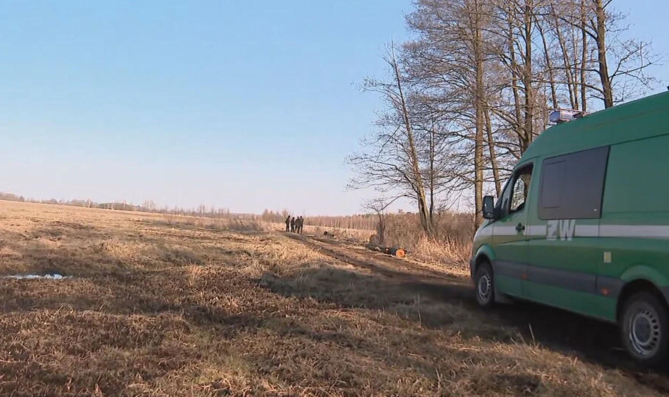 Zielony policyjny bus stoi zaparkowany na wiejskiej drodze przy suchym polu; w oddali grupa ludzi zbliża się w kierunku pojazdu, a w tle widoczne są bezlistne drzewa.